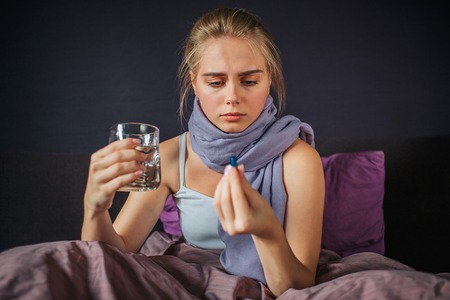 Concentrated young woman sits on bed and looks at medicine she holds with fingers. There is glass of water in another hand.の写真素材