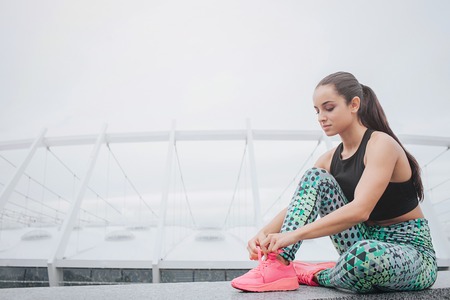 Pretty and beautiful young woman sitting and having rest. She ties laces on her sports shoes. Model smiles. She is positive. Young woman sits near big stadium.の写真素材