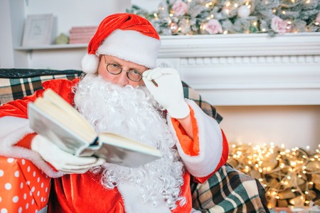 Serious and concentrated man in santa claus costume reads book. He holds hand on glasses. Guy pays attention to book.の写真素材