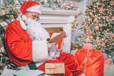 Serious and thoughtful man in Christmas suite sits and reads letters. There are other besides him on table. Man sits at fireplace and Christmas tree.の写真素材