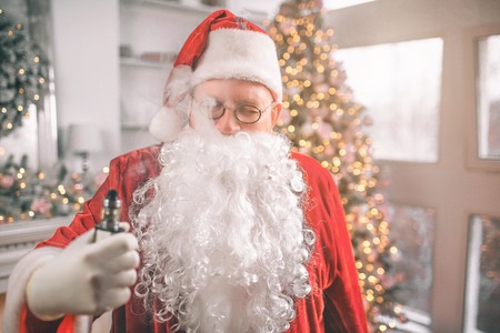 Relaxed man in Santa Claus costume stands and holds vape in hand. Smoke comes out of his mouth. He is relaxed.の写真素材