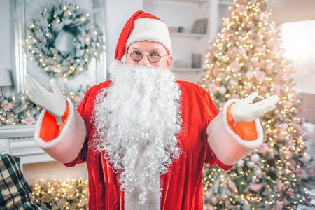 Happy Santa Claus stands and looks on camea. He poses with hands aside of body. Man smiles. There are festive Christmas tree and fireplace behind him.の写真素材