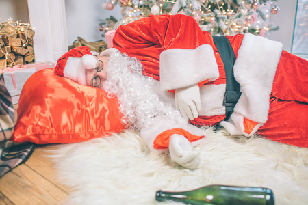 Drunk Santa Claus lying on floor and sleeping. His bag is under head. Bottle lying on carpet.の写真素材