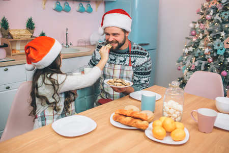 Small girl sits at table and feeds her dad with cookie. He smiles and looks at her. Young man bites cookie. They wear festive clothes and red hats.の写真素材