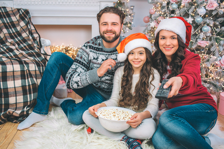 Nice and cheerful family sitting on floor and watch straight. They smile. Family is in decorated room. Young woman holds remote control and push button on it. They wear festive clothes.の写真素材