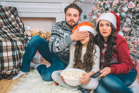 Scared parents look straight on camera. Father coveres eyes of his daughter with hand. They watch together. Girl holds bowl of popcorn. She sits between parents.の写真素材
