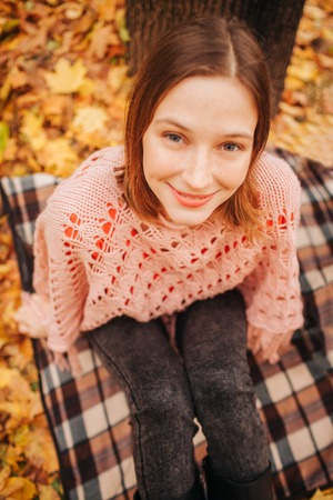 Portrait of young woman sitting alone in park. It is autumn outside. She looks on camera and smile.の写真素材