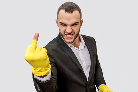 Angry and rude young man in suit stands and look on camera. He shows fuck. His hands are in yellow gloves. Glove on middle finger is wrapped up. Isolated on white background.の写真素材
