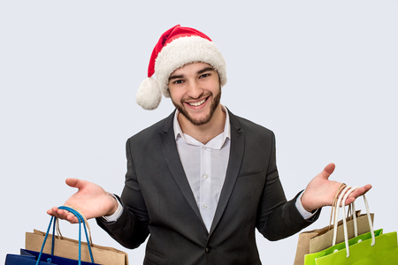 Cheerful and positive young man smiles on camera. He wears Christmas hat and black suit. Young man has shopping bags in both hands. Isolated on white background.の写真素材