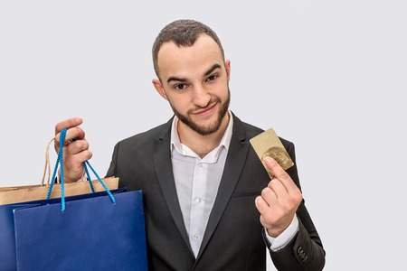 Nice and confident young man in suit stands and shows credit card. He looks straight. Young man has shopping bags in hand. Isolated on white background.の写真素材