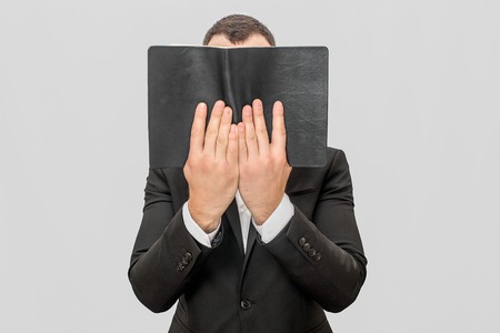 Young businessman coveres face with book in dark cover. He hold it with both hands. Guy weas suit. Isolated on white background.の写真素材
