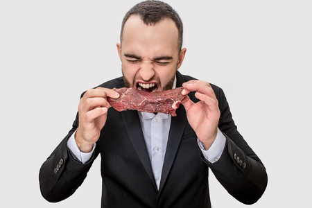 Picture of young businessman screaming and yelling. He holds piece of raw meat. Guy is going to bite it. Isolated on white background.の写真素材