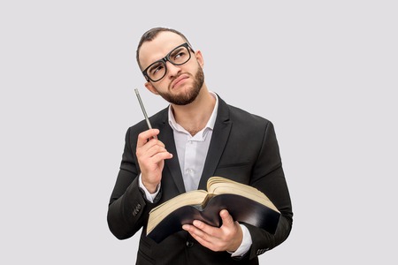 Thoughtful and serious young man holds book and pen. He looks up though glasses. Young man wears...