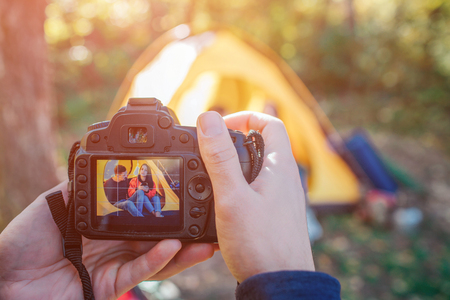 Man holding camera that take picture of couple. They sit in tent. People pose. She holds thermocup. He looks at her.の写真素材