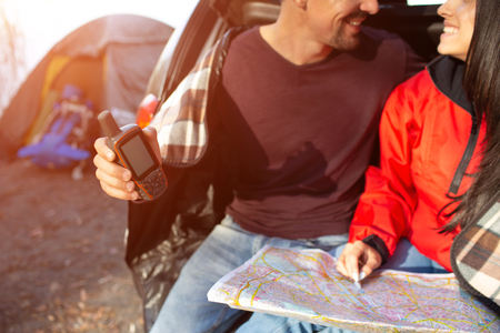 Cut view of lovely couple sitting together in trunk and smile. He holds satellite phone. She has map. People are close to tent.の写真素材