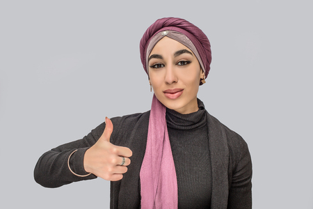 Young arabian woman stand and look on camera. She holds big thumb up. She wears hijab. Isolated on grey background.の写真素材