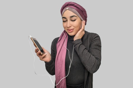 Cheerful young arabian woman hold phone. She listen to music through headphones. Model keep eyes closed. Isolated on grey background.の写真素材