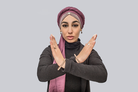 Serious young arabian woman stand and hold hands in crpss position. She looks on camera. Model wears hijab and black dress. Isolated on grey background.の写真素材