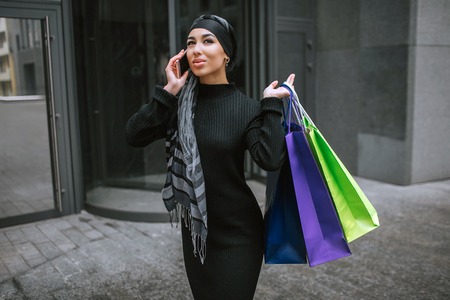 Modern and fashioned young arabian woman walks and talks on phone. She cary shopping bags behind...