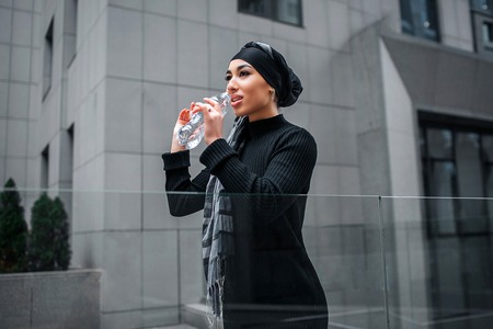 Attractive arabian woman drinks water from bottle. She looks straight forward. Woman stands at edge of glass railings.の写真素材