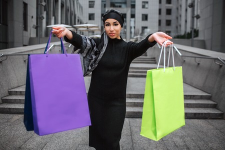 Confident young arabian woman hold colorful shopping bag and look at camera. She smiles a bit. Model wears black dress and hijab.の写真素材