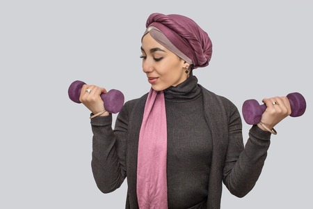 Strong young arabian woman exercising with dumbbells. She looks at one of it. She wears hijab. Isolated on grey background.の写真素材