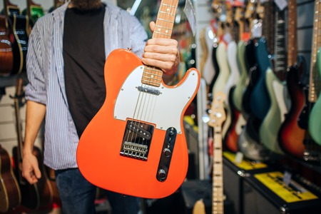 Cut view of man in guitar shop holding electric instrument in hand. He show it to camera. Man is...