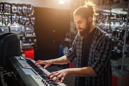 Serious and concenrated young hipster sit and play on keyboard. He is alone in room. It is sunny...