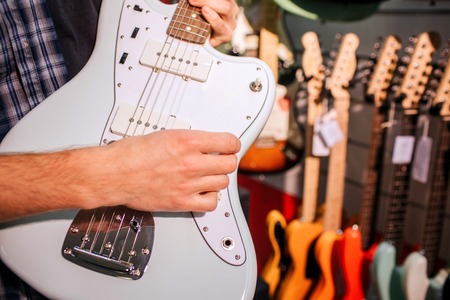 Close up of man holding light-blue guitar in hands. He plays on it and touches its strings. Many colorful electronic guitars hanging on wall behind.の写真素材