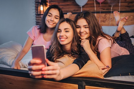 Three young models taking selfie. They pose and smile. Gils are in room. One of them hold phone and take picture on it. Young women are cheerful.の写真素材