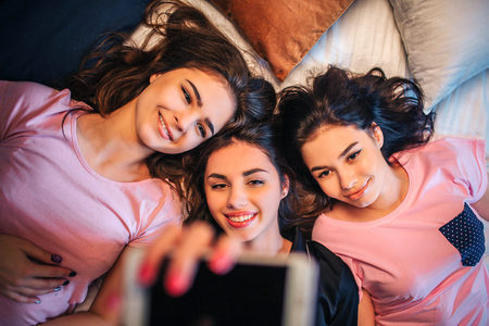 Three young women lying on bed. Girl in middle hold phone in hand. They take selfie and smile to...