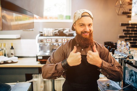 Happy young barista stand and show big thumb up on both hands. He look on camer. Guy stand in kitchen. Light comes out of window.の写真素材