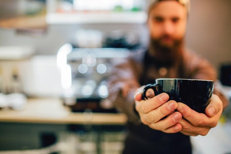 Close up f dark cup. Guy hold it with both hands. He stands in kitchen. Coffee machine is behind him.の写真素材