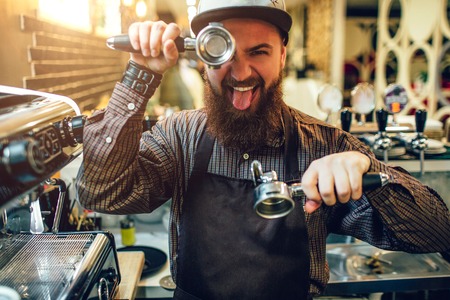Funny young bearded man covering one eye with cezve and showing tongue on camera. He stands in kitchen at coffee machine.の写真素材