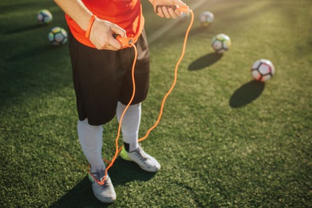 Cut view of man standing on green lawn outside. He hold orange jump rope in hands. Five balls lying behind. Sun is shining.の写真素材