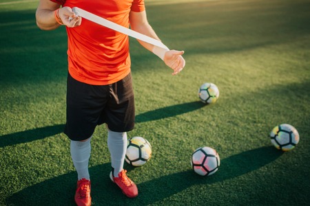 Cut view of football player holding bandage in hands. He stretch it. Balls are on lawn. He stand there as well.の写真素材