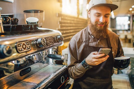 Cheeerful young man stand at coffee machine and look at phone. He smiles. Guy hold cup of coffee as well. It is light inside.の写真素材