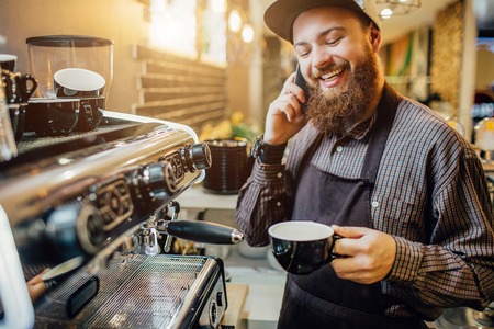 Nice bearded barista stands at coffee machine and talks on phone. He smiles. Guy hold cup of coffee in hands.の写真素材