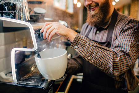 Picture of young barista pouring some coffee into huge white cup. He does it with smile and pleasure. Guy stand at coffee machine in kitchen.の写真素材