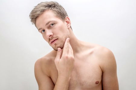 Handsome young man looking straight. He touches face. Guy is shirtless. Isolated on white background.の写真素材