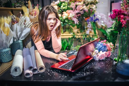 Picture of young female florist at table. She look at laptop and wonder. Paper rolls are on table....