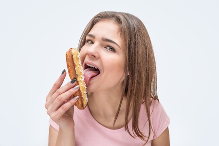 Cheerful young woman look on camera and lick eclair. Isolated on grey background.