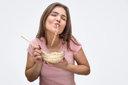 Joyful young woman eat vermicelli. She hold bow with it and fork in hands. Isolated on white background.の写真素材