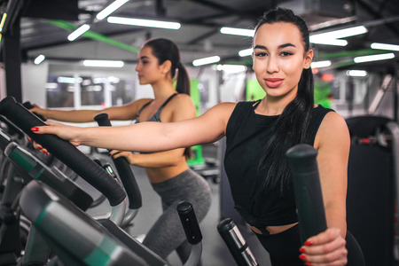 Nice young asian woman look on camera. She works on cross-trainer simulator. Another sexy young...