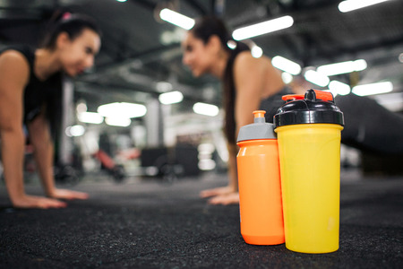 Orange and yellow water bottles stand in front of picture. Asian and european women stand in plank pose behind. They look at each other and smile. Models work in gym.の写真素材