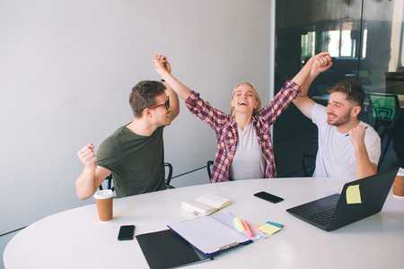 Very happy young coworkers cheering. Guy hold blonde model hands. All of them smile. They look at each other. People are very positive. They in one roomの写真素材