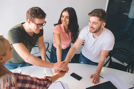 For cheerful and happy coworkers stand around white table and look at each other. They hold hands...