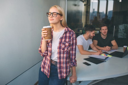 Smart young blonde woman wear glasses. She stand and hold cup of coffee. She enjoyes. Two young men sit behind her and work. They talk between themselves.の写真素材