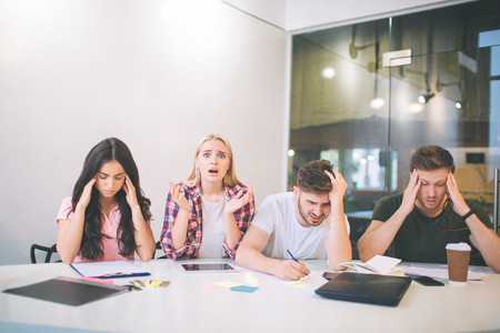 Amazed and upset young blonde woman look on camera. She scream. Other people trying to concentrate. They do brainstorming. All of them sit together in white room.の写真素材