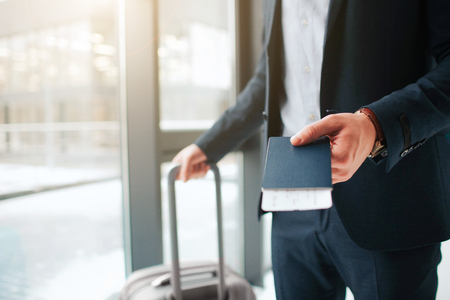 Cut view of man in suit stand at window in airport. He hold passport with ticket in it and suitcase with another hand.の写真素材